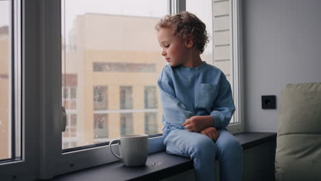 caucasian little boy is looking out window from apartment on high floor watching snow in winter day
