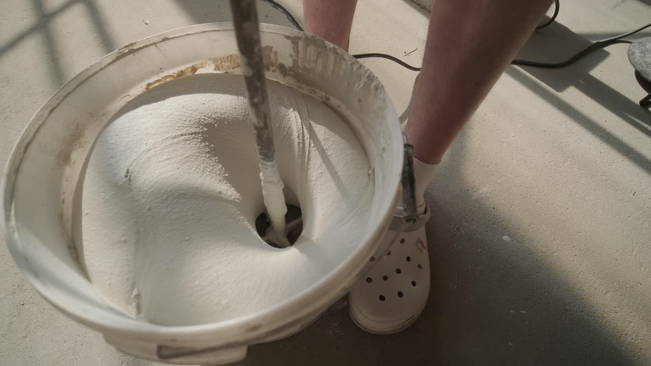 Close-up of a construction worker's legs beside a bucket as he mixes a batch of white plaster with a mixing tool, casting shadows on the ground