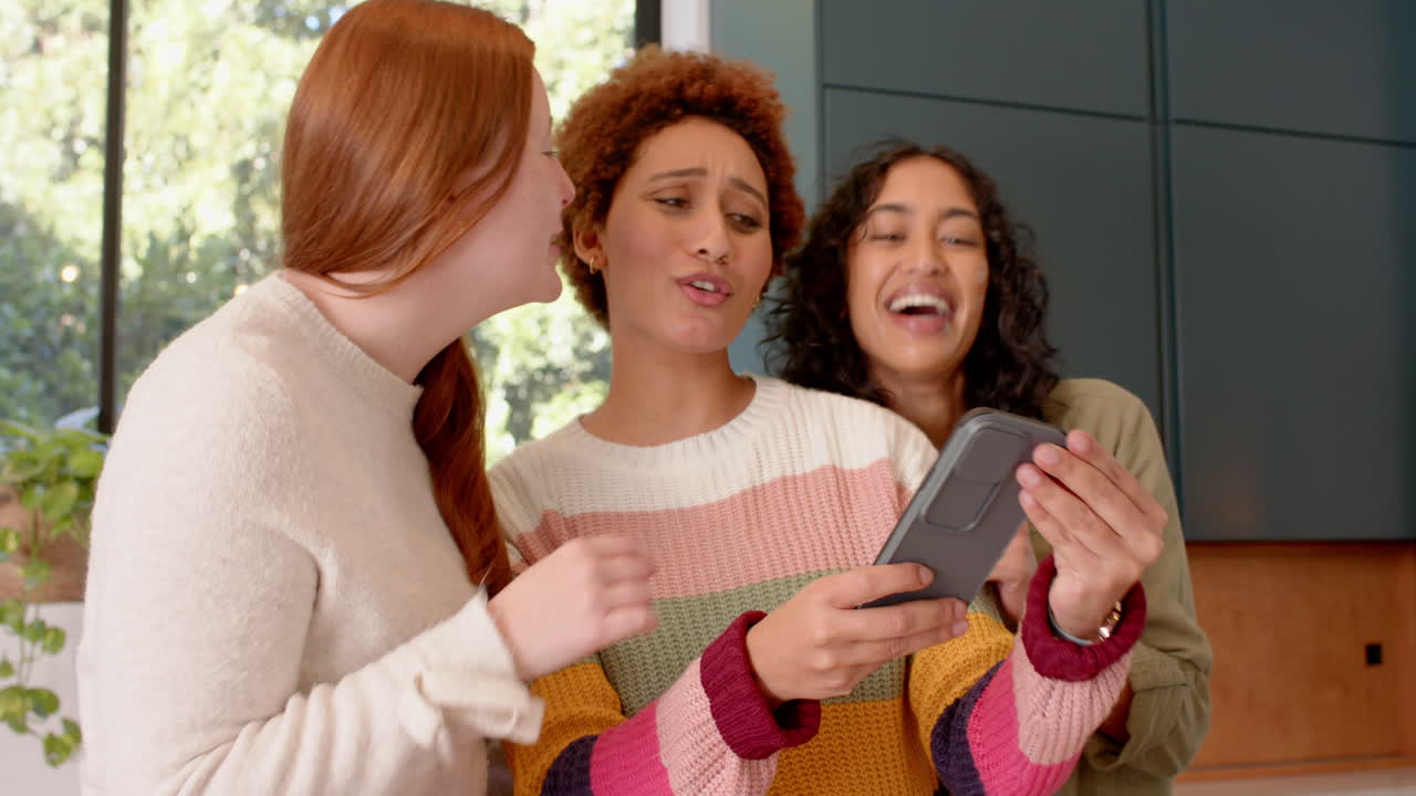Looking at smartphone, smiling female friends enjoying time together indoors