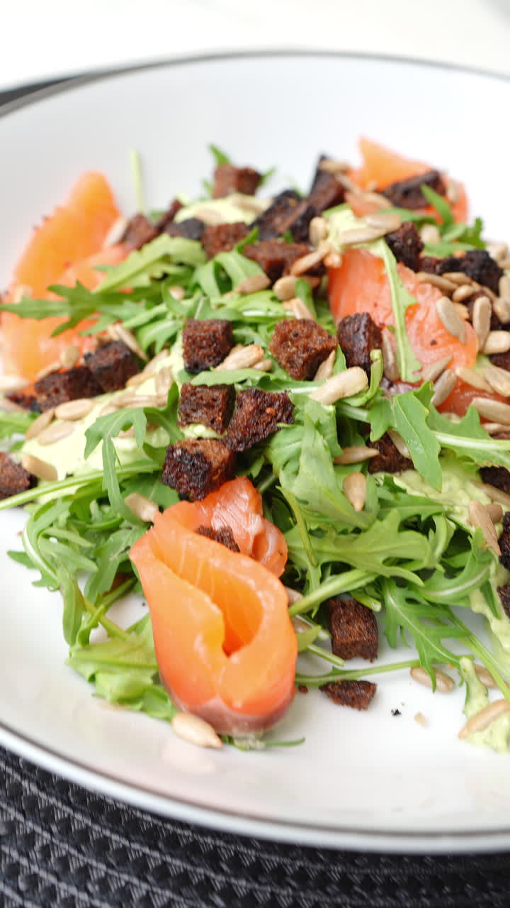 Vertical footage Salmon salad spinning with arugula, crispy croutons, sunflower seeds, creamy avocado sauce on white ceramic plate, dark background