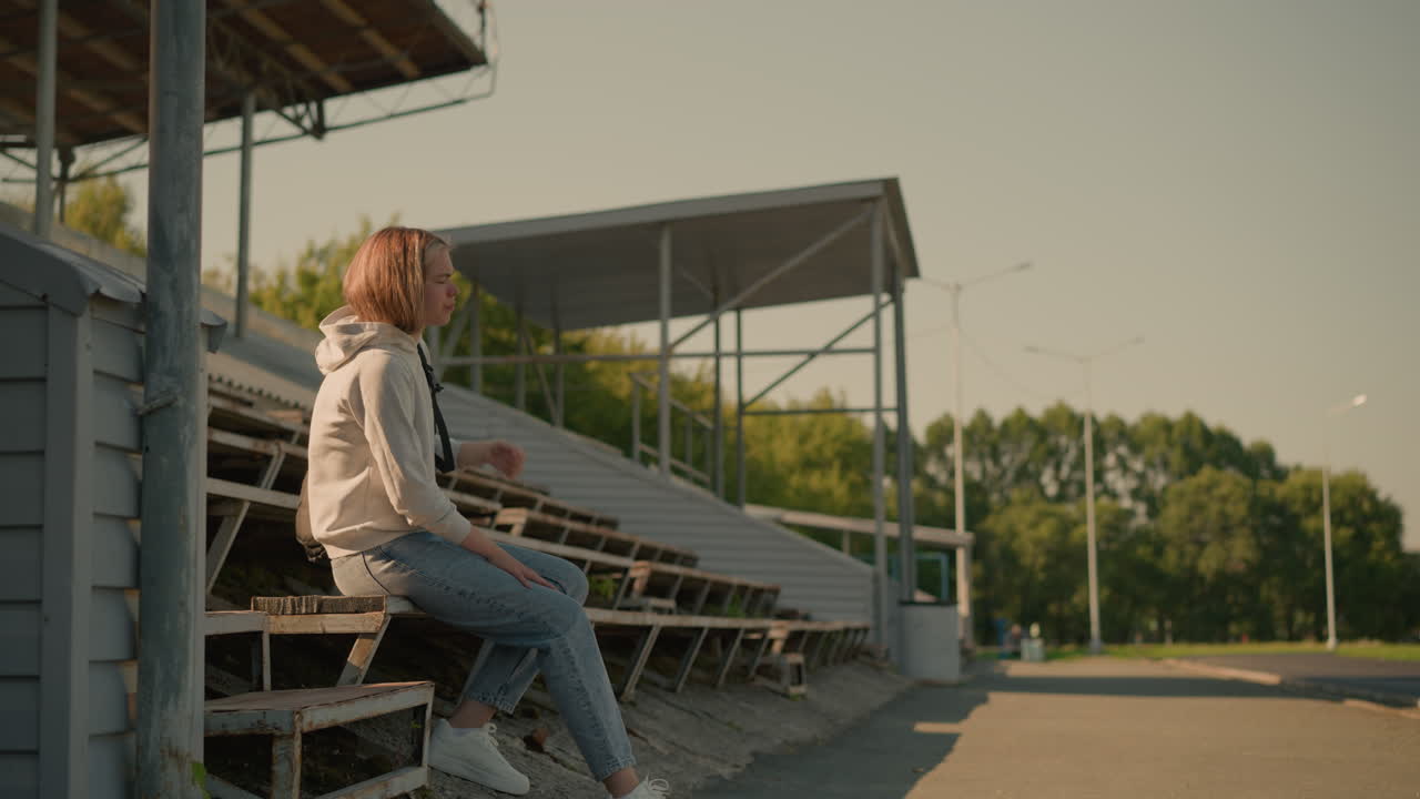 검은 가방을 입은 후디를 입은 젊은 여성이 오른쪽 다리를 앞두고 농촌 스타디움 관중석으로 걸어가고, 약간 앞으로 기울어 그녀의 자세를 조정합니다.