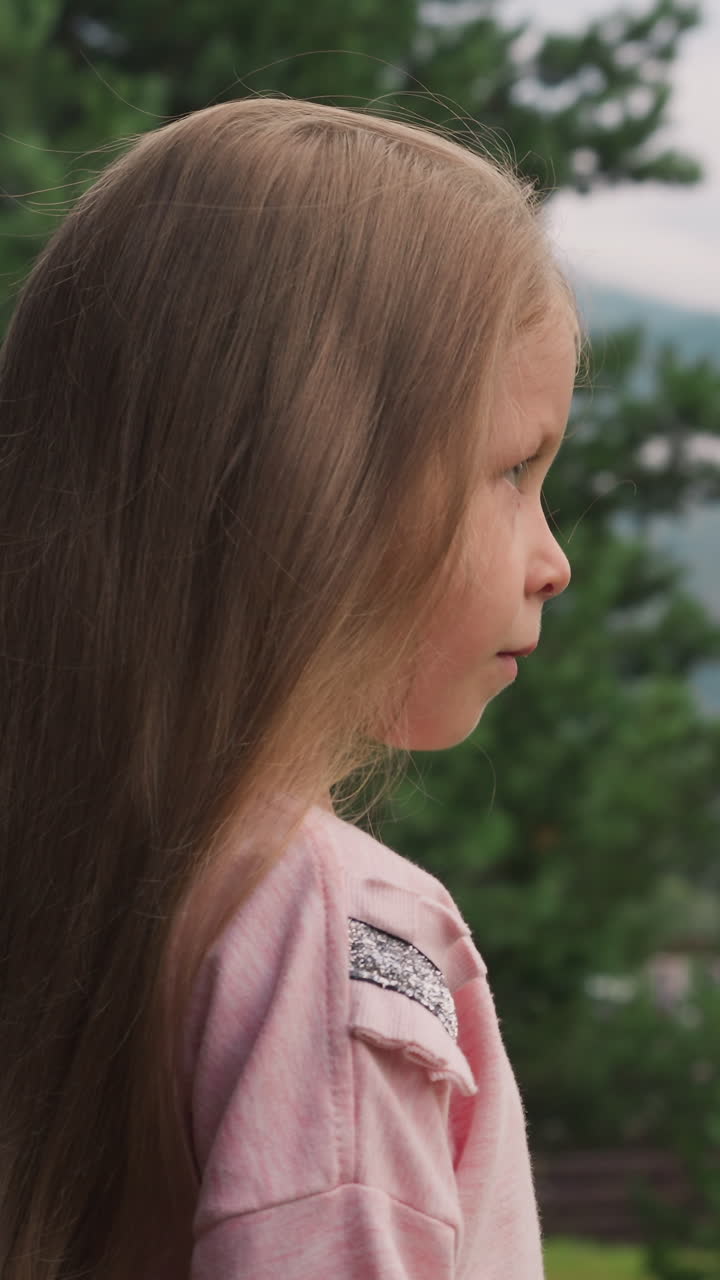 tranquila niña con cabello rubio largo y suelto se encuentra cerca del pino contra grandes montañas distantes en un día nublado vista lateral cercana cámara lenta