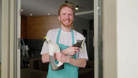 Portrait-of-a-confident-blond-man-with-a-beard-in-a-white-T-shirt-and-with-a-blue-apron-a-cleaner-holds-in-his-hands-two-vacuum-cleaners-for-windows-during-his-cleaning-in-a-modern-apartment-on-call-from-a-cleaning-company