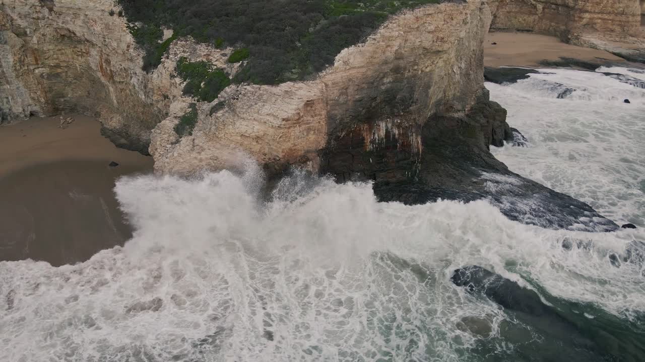 A slow motion drone shot circling a hidden sea arch over turquoise Pacific ocean saltwater with waves crashing against cliffs near Santa Cruz off the historic Highway 1 with yellow California poppies