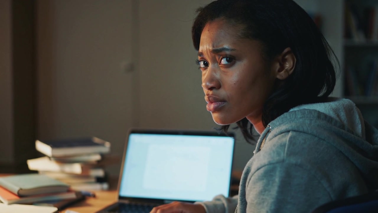 Pausing typing, student in grey hoodie turning head at desk, noticing laptop lamp and stacked books