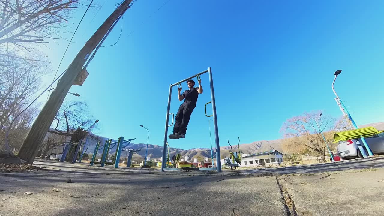 Low-angle shot of a man in black outfit performing pull-ups in a city outdoor gym under clear blue sky