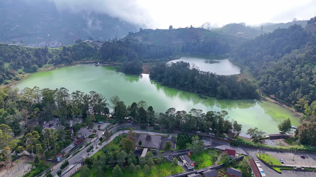 Aerial footage of a volcanic lake with emerald-colored water, located in a lush mountain valley. Misty forest, calm atmosphere, and dramatic highland landscape. Telaga Warna Lake, Dieng, Indonesia