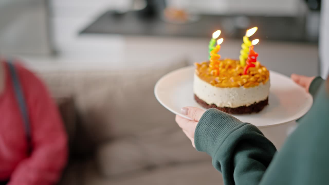 sobre su hombro una chica morena con una chaqueta verde trae un pequeño pastel con velas para su padre en honor a su cumpleaños. un hombre mayor feliz con cabello gris y una barba exuberante en una camisa rosa sopla velas y agradece a su hija adulta por sus deseos de cumpleaños en un apartamento moderno
