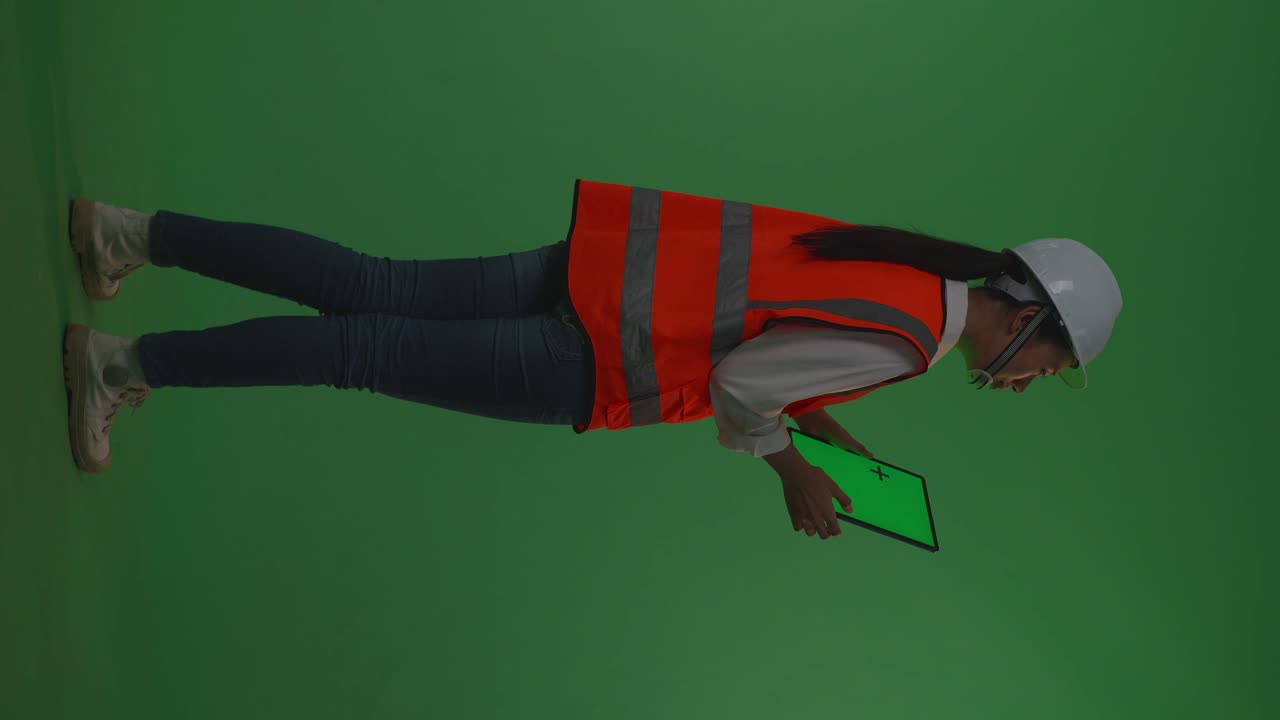 vista trasera de cuerpo entero ingeniera asiática con casco de seguridad trabajando en una tableta de pantalla verde y mirando a su alrededor mientras está de pie en el estudio de fondo de pantalla verde, durante la puesta o salida del sol