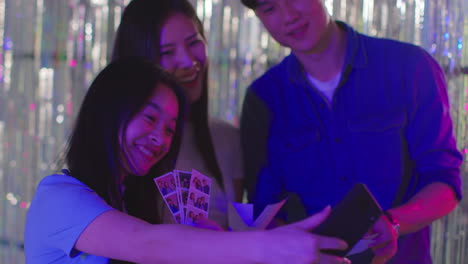 Group-Of-Friends-Taking-Selfie-Having-Fun-Looking-At-Prints-From-Photo-Booth-In-Nightclub-Or-Funfair