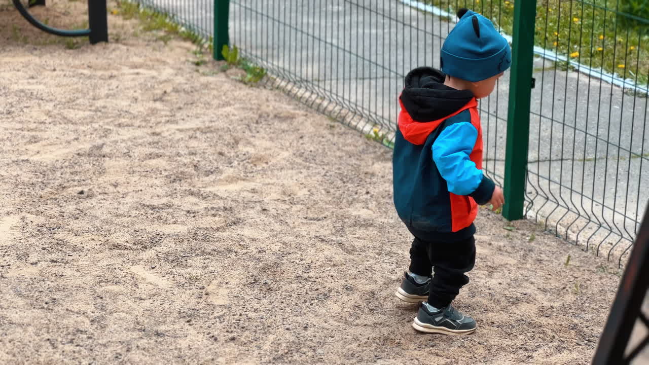 Caucasian child plays with sand on the playground. Kid takes the handfuls of sand and quickly throws through the fence.