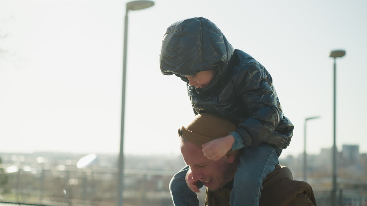 아들을 어깨에 고 있는 아버지의 클로즈업, 아들이 아버지의 후드와 함께 놀고 있는 모습, 아버지는 갈색 재을 입고, 아들은 검은색 재를 입고, 도시 풍경이 흐릿하게 보인다.