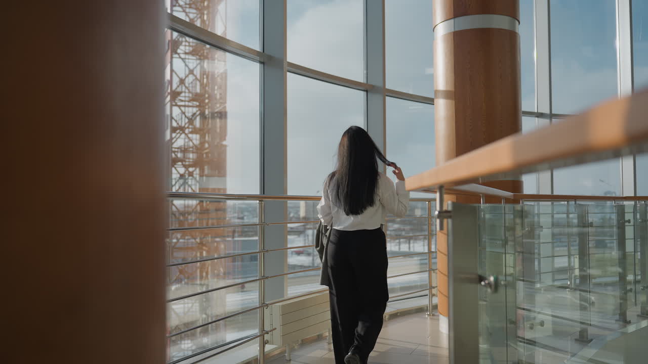 Back view of woman in white shirt walking inside modern mall holding jacket and touching her hair with glass windows in background revealing blurred snowy urban scenery