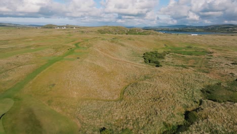 Drone-flyover-Ireland-golf-course,-views-of-rolling-hills,-fairways,-bunkers,-ocean-and-mountains-in-background