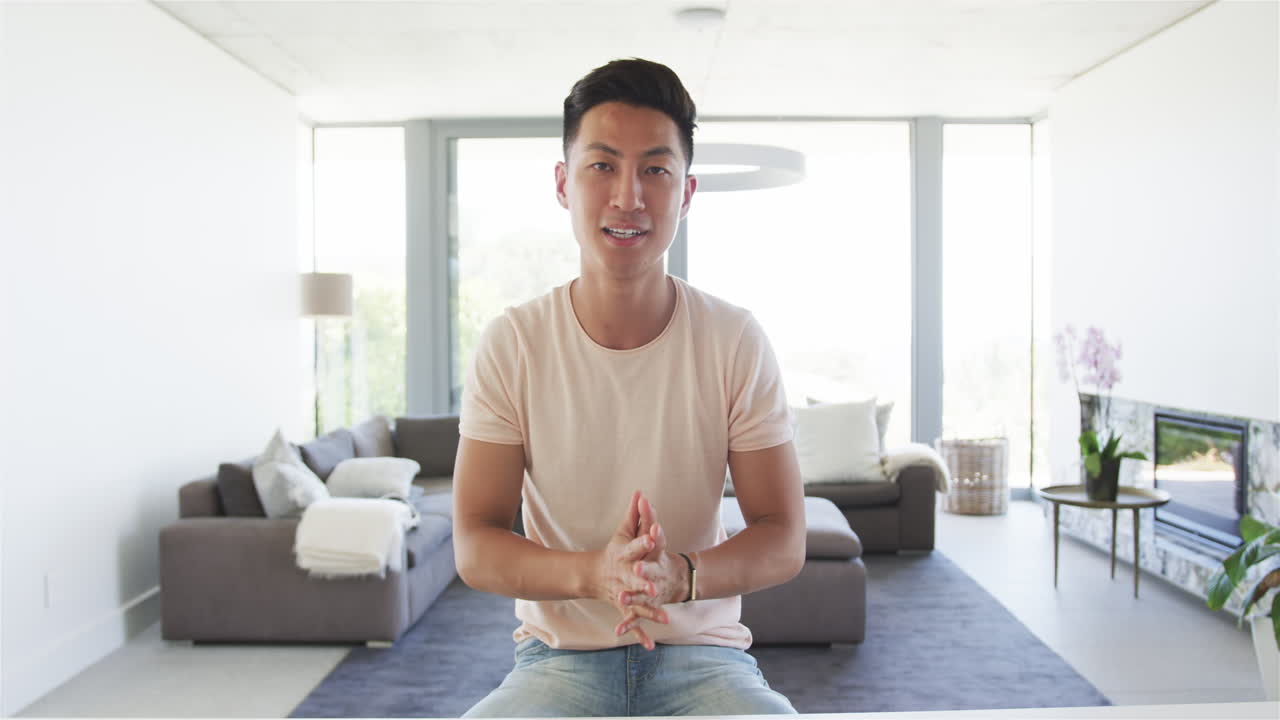 젊은 아시아인 남자가 현대적인 거실에서 비디오 통화에 무관하게 앉아 있습니다.