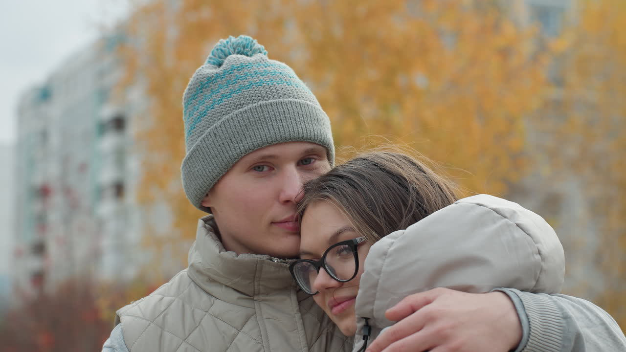 Close up of two friends embracing warmly in identical winter outfits outdoors with soft view of golden autumn trees and buildings in background