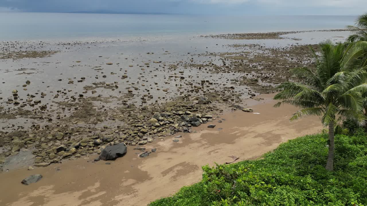 A drop-down drone shot over a tropical shoreline at low tide, revealing sandy beach, rocky shallows, palm trees, and calm ocean under a cloudy sky near Mauban Port, Quezon Province Philippines