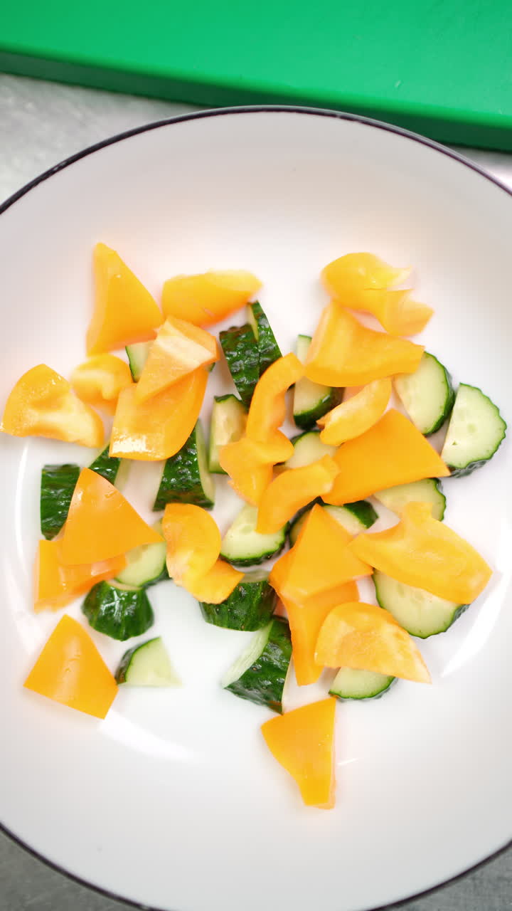 Vertical footage Professional chef wearing black protective gloves chopping orange bell peppers, adding ingredients to white mixing bowl with sliced cucumbers on green wooden cutting board