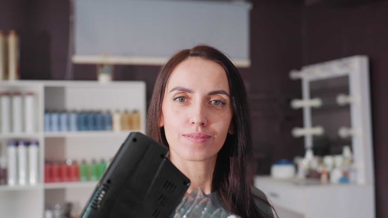 Estilista de peluquería probando un secador de aire caliente cerca de la cámara. Una estilista blanca muestra el flujo de aire y los ajustes mientras examina la boquilla y el mango. El moderno fondo de estantería de productos transmite una reseña profesional.