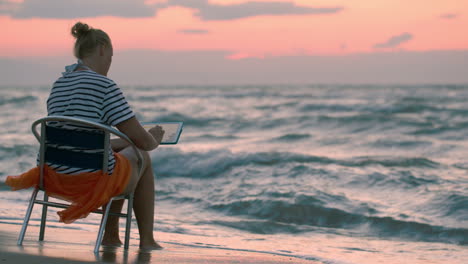 Woman-with-pad-sitting-on-chair-by-rough-sea