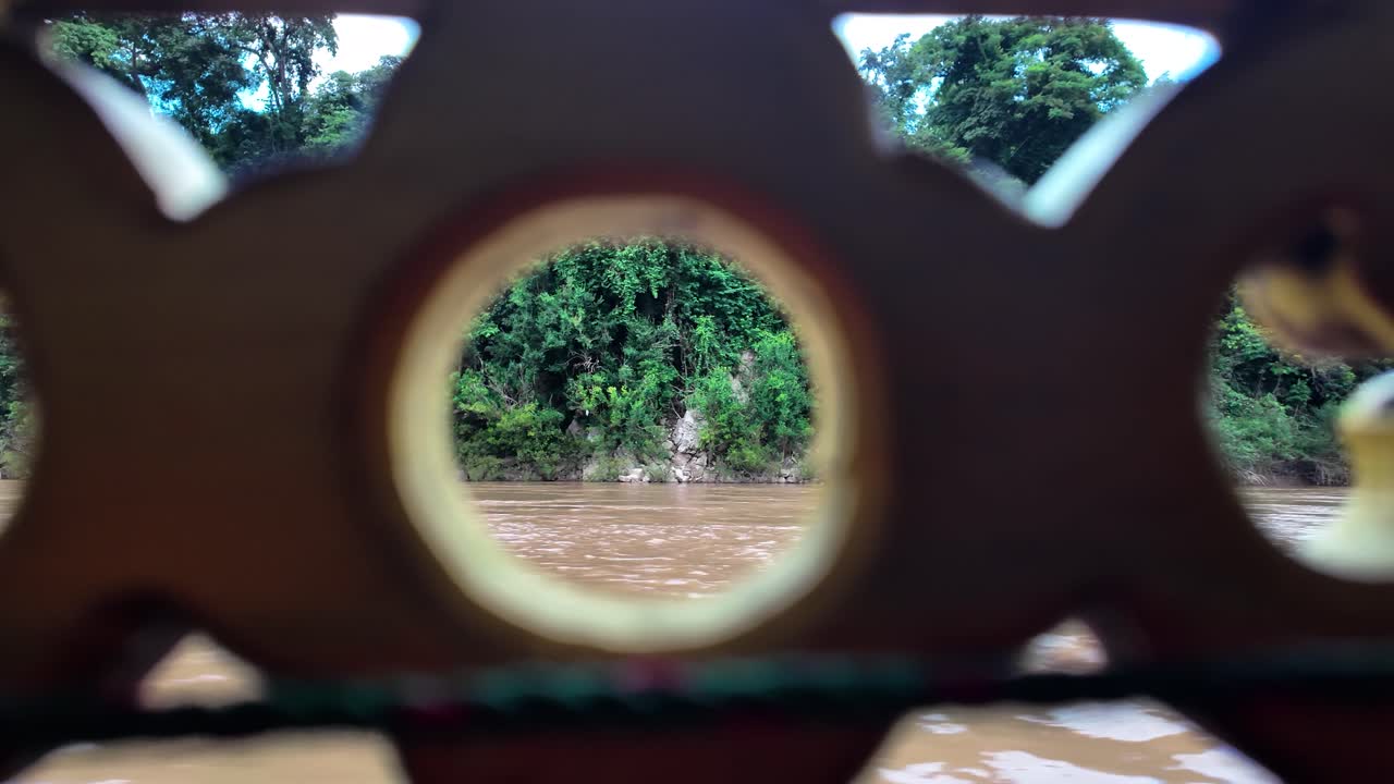A view of the Mekong River and forest riverbank in Laos seen through a carved wood railing on a slow boat