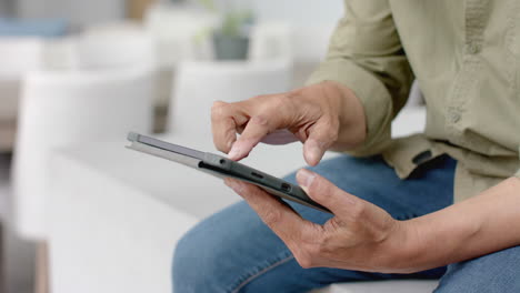 Midsection-of-senior-biracial-man-sitting-on-table-using-tablet-at-home,-slow-motion