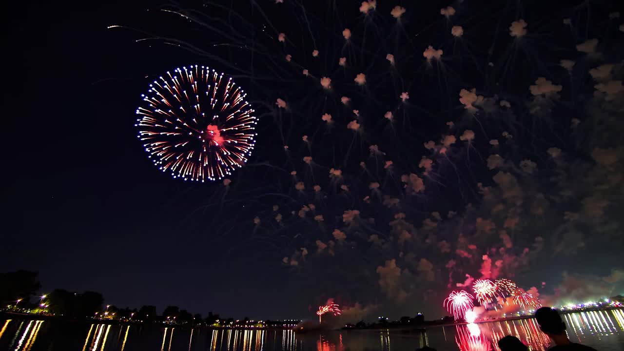 A wide-angle video captures vibrant fireworks exploding over a serene lake at night