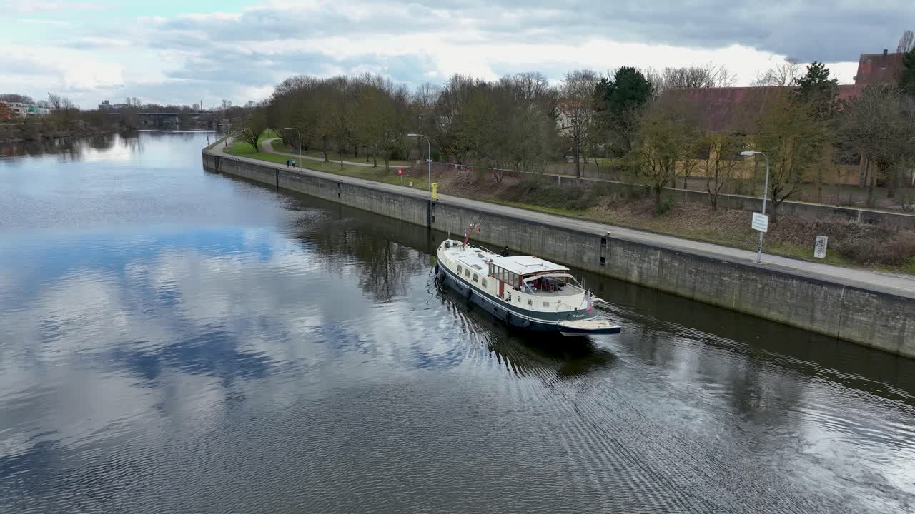 Aerial drone footage follows a charming houseboat as it sails along a serene city canal in Germany. The calm water beautifully reflects the sky