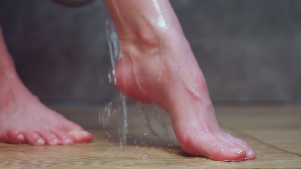 close up partial view of woman raising left leg under gentle stream of water in modern tiled shower, wet foot pressing onto wooden floor, droplets glistening on skin