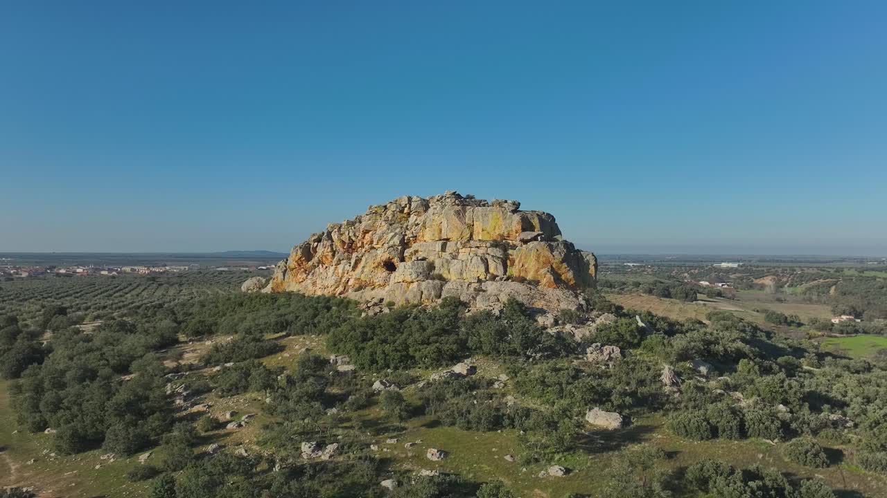Spectacular drone flight above an olive field highlighting a rocky hill rising from the plain. Its vivid colors yellow, orange, green,surround a striking hollow that forms a cave entrance