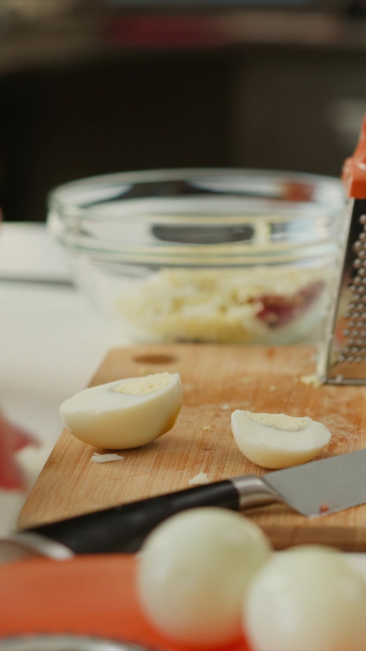Mano di donna affetta con cura un uovo sodo su un tagliere di legno, mettendo ogni metà in una ciotola di vetro. La scena presenta dettagli ravvicinati di un coltello e metà cipolle sullo sfondo sfocato.