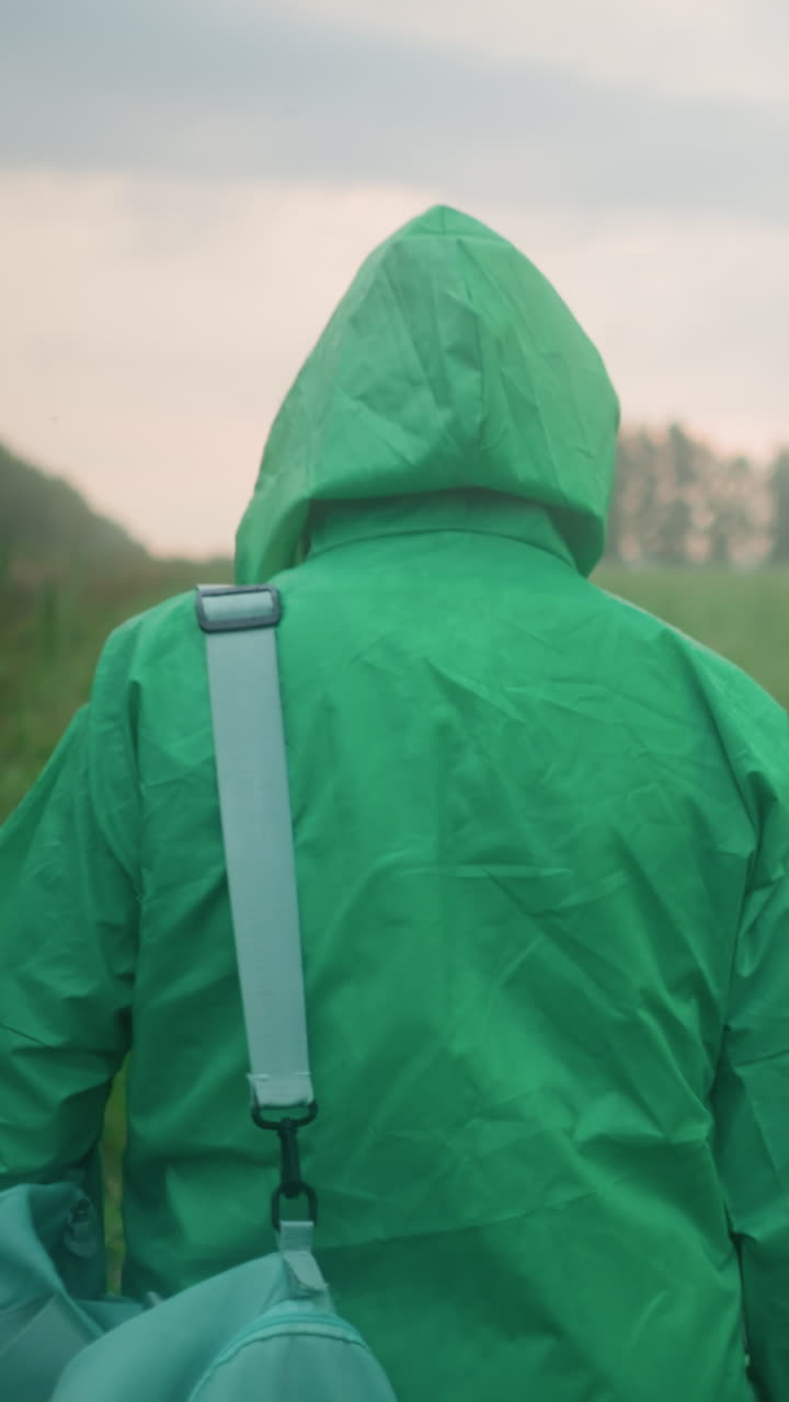 은 하늘 아래 산란한 덤불과 나무로 둘러싸인 안개가 인 초록색 을 고 가방과 우산을 들고 있는 레인코트를 입은 개인의 뒷면