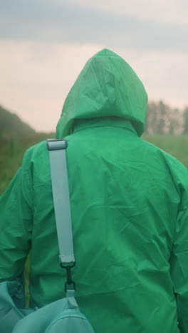 vista trasera de un individuo en impermeable que lleva una bolsa y un paraguas caminando a través de un campo verde y brumoso, enmarcado por arbustos y árboles esparcidos bajo un cielo nublado con un sol lejano mirando a traves de las nubes