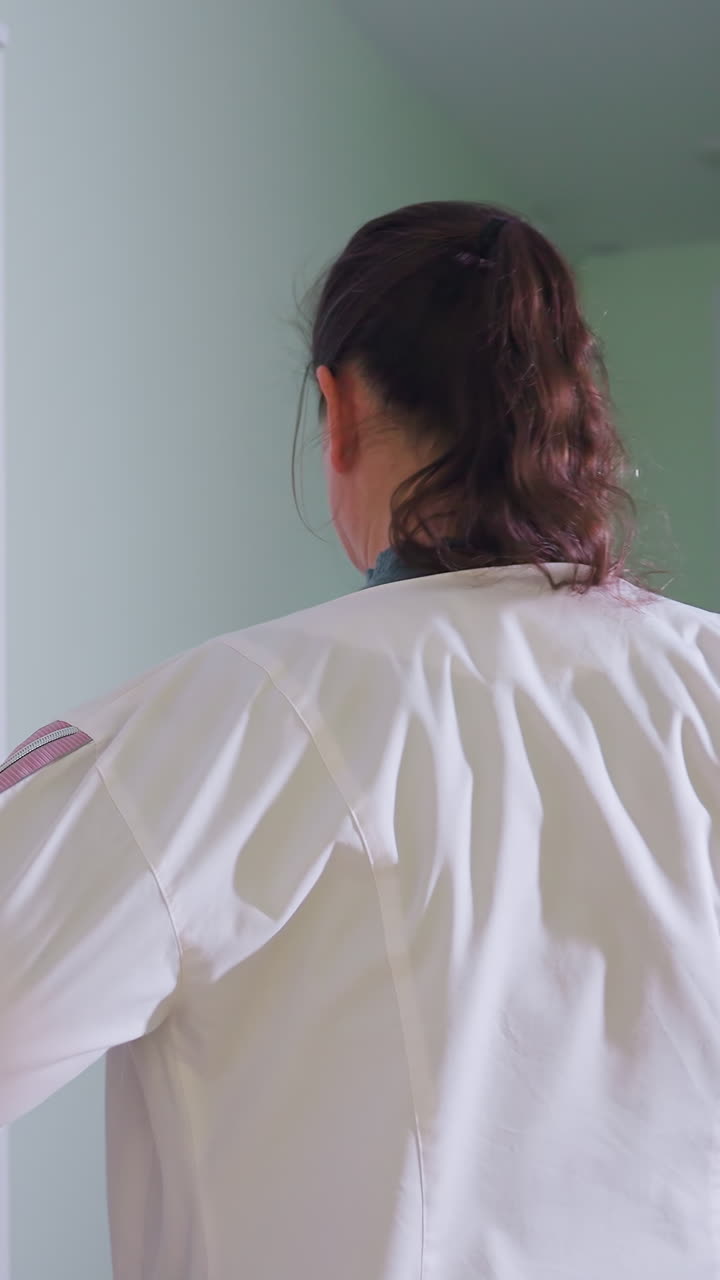 작은 진료실에서 진료를 받기 전에 흰색 코트를 준비하고, 소매를 다듬고, 포니테일을 집어넣고, 자신감 있는 자세로 미소를 짓는 백인 여성, 배경에는 초음파 모니터와 침대가 있으며 따뜻한 분위기