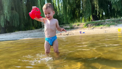 lovely toddler stands in the river to his knees. baby boy fills his watering can and pours some water on himself.