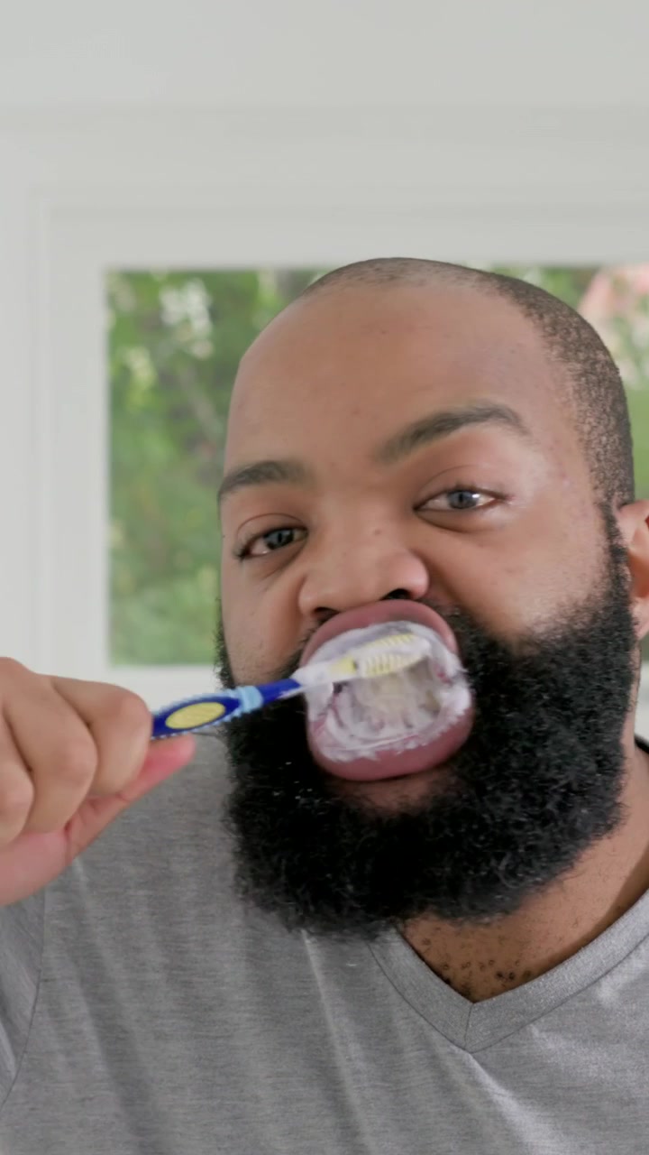 Vertical video: Bringing brush with foam, man in gray Vneck brushing teeth at bathroom sink