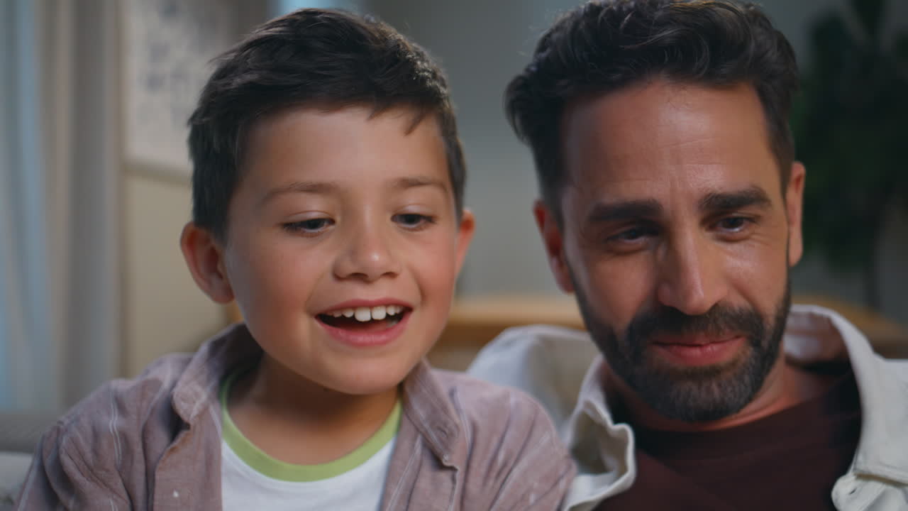 Positive dad son watching movie on computer sitting cozy couch at home closeup