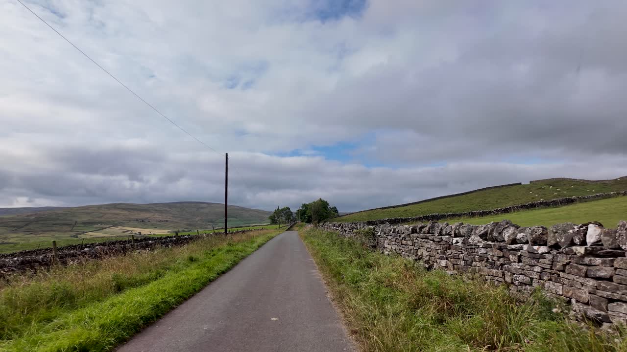 A narrow country road winding through vast green landscape with stone walls and rolling hills