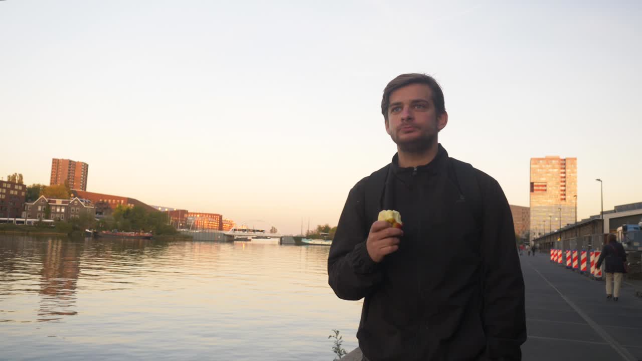 vídeo de un hombre caminando por un muelle en amsterdam y comiendo una manzana durante el atardecer