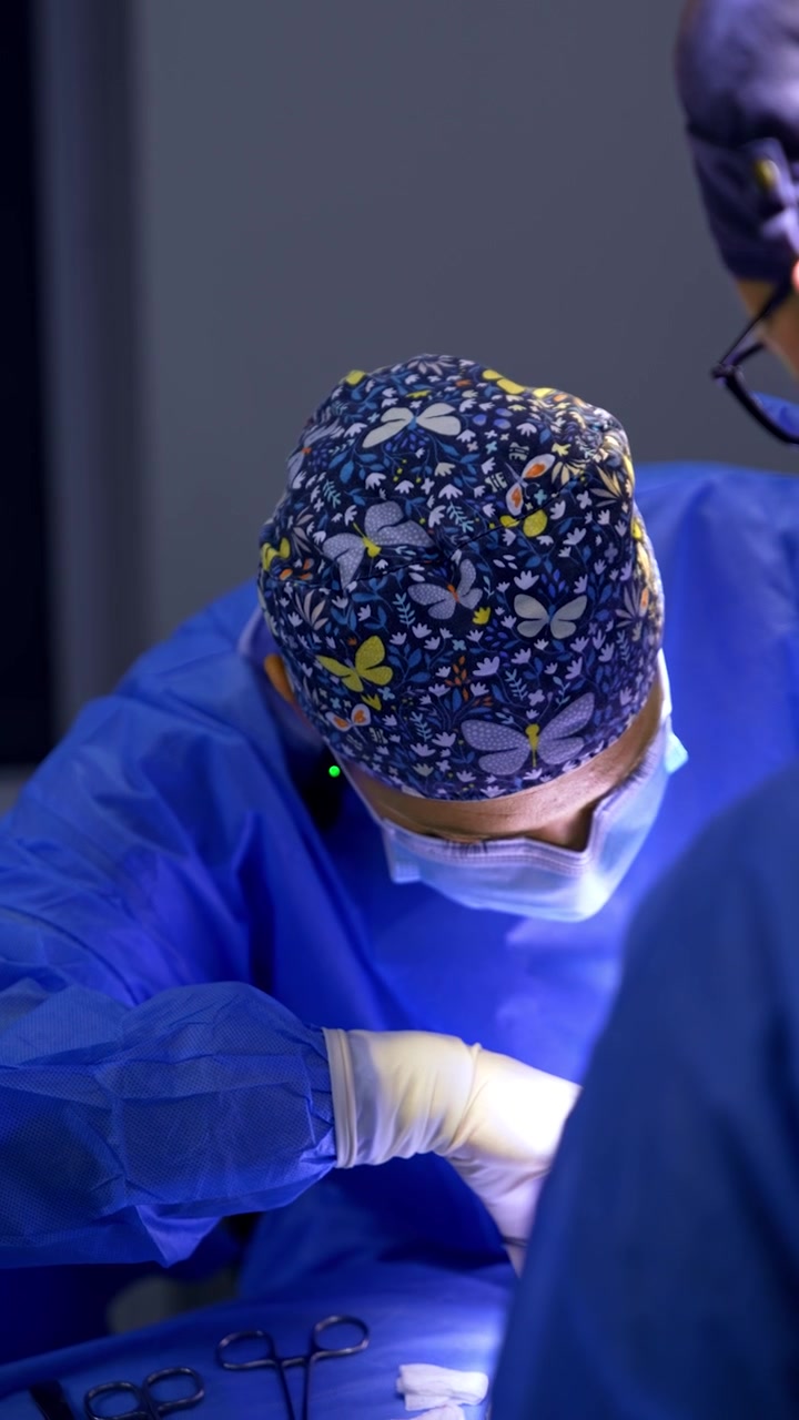 Cirujana con gorro de mariposas inclinada sobre el paciente. La doctora realiza la operación. Video vertical.