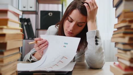 Exhausted-business-woman-looking-at-documents-reports-stacks-of-books-while-working-hard-at-office