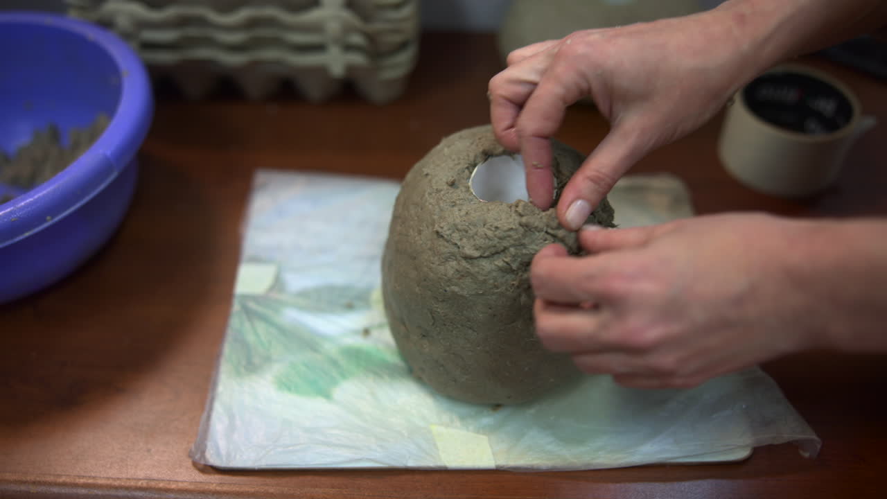 Hands Shaping Mud Dome Around White Candle, Artisan At Workshop Molding Textured Surface On Towelcovered Table. Media