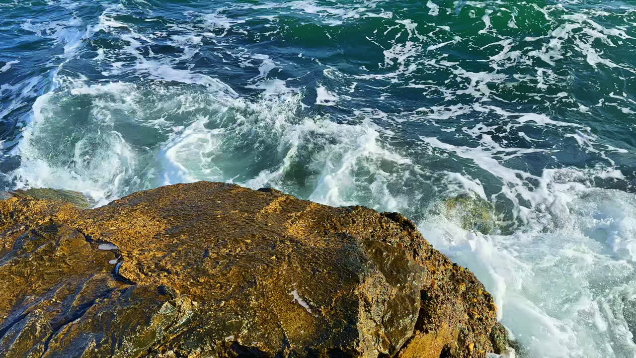 Le onde si infrangono contro le rocce sulla riva a mezzogiorno in un'area costiera con acqua limpida e luce solare.