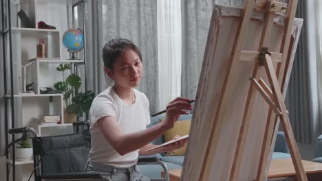 medium shot side view of an asian artist girl in wheelchair holding paintbrush mixed colour thinking and painting on the canvas