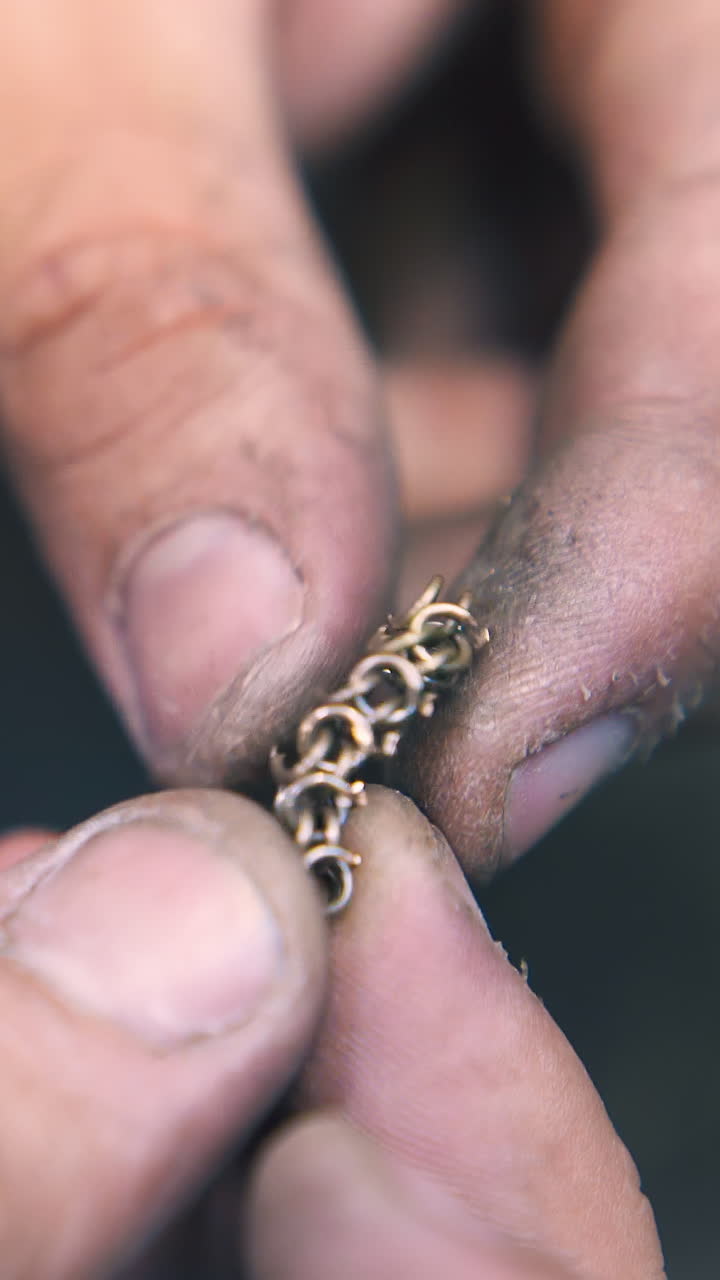 CU: skilled jeweler connects little gold link and looks at chain at workplace in light craft workshop extreme closeup