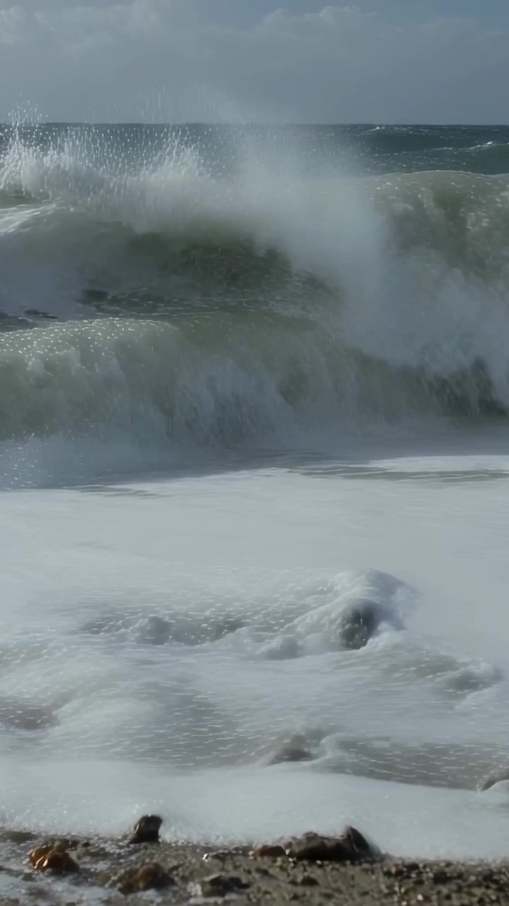 Video verticale: Mareggiata in arrivo che spinge onde che si infrangono e si rompono su una spiaggia di ciottoli, rocce spumeggianti.