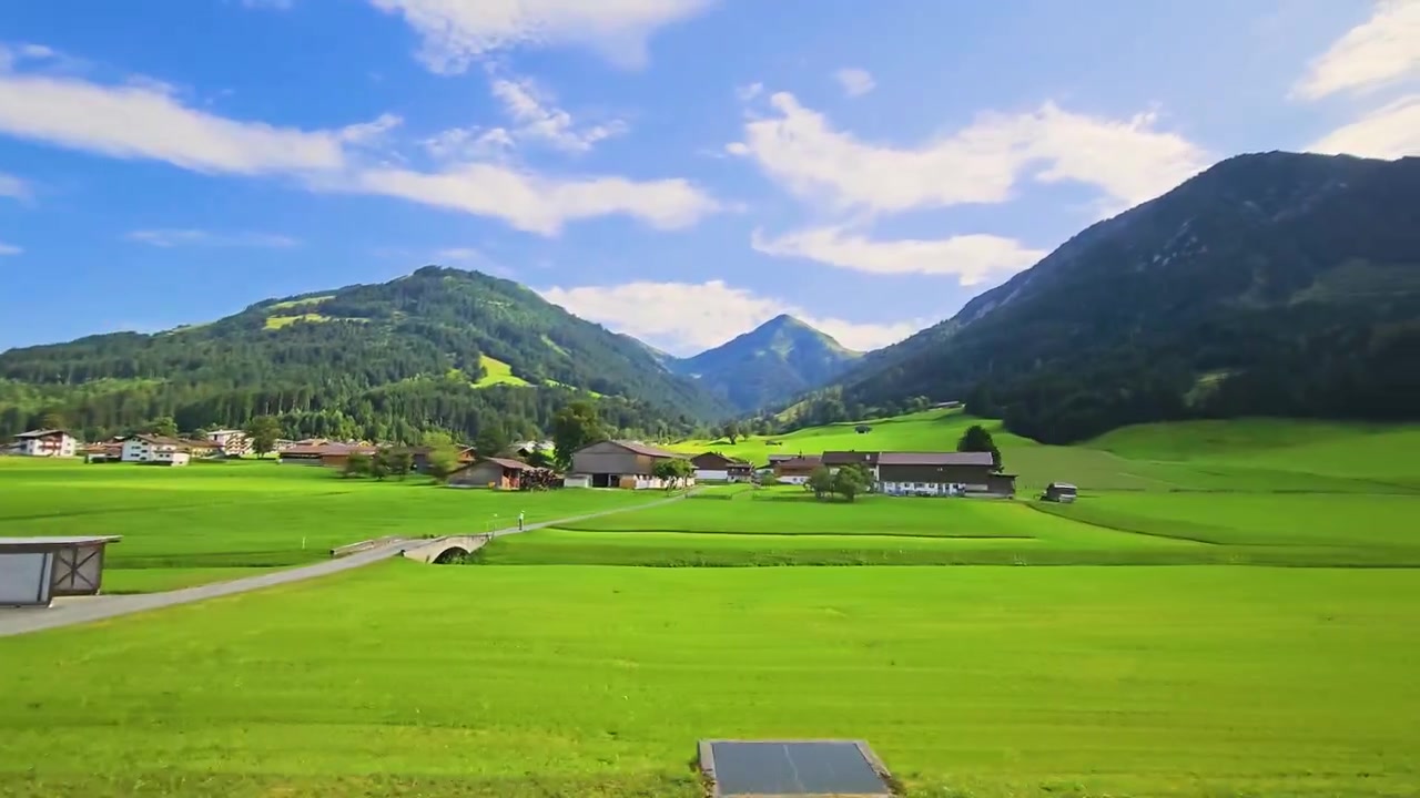 Scena di viaggio in treno con campi d'erba luminosi, tetti di villaggi, pendii boscosi e cime montuose, filmata da un finestrino mentre si attraversa le Alpi in un paesaggio rurale pacifico.