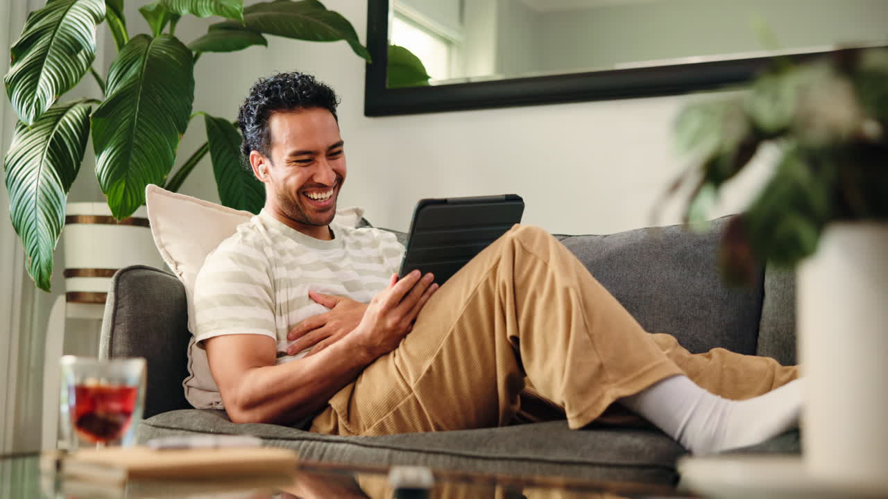 Happy Man Laughing While Using Digital Tablet on Couch at Home