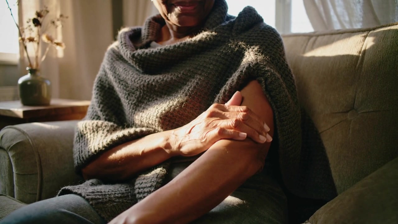 Sentendo la luce del sole, una donna anziana si gira sul divano vicino alla finestra, tirando la manica del poncho e accarezzandosi l'avambraccio.