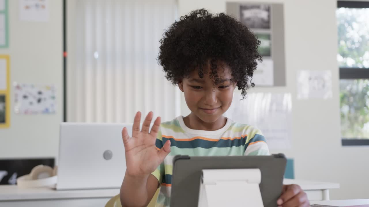 African American boy smiling at tablet on white stand at desk, spotting classmate and waving